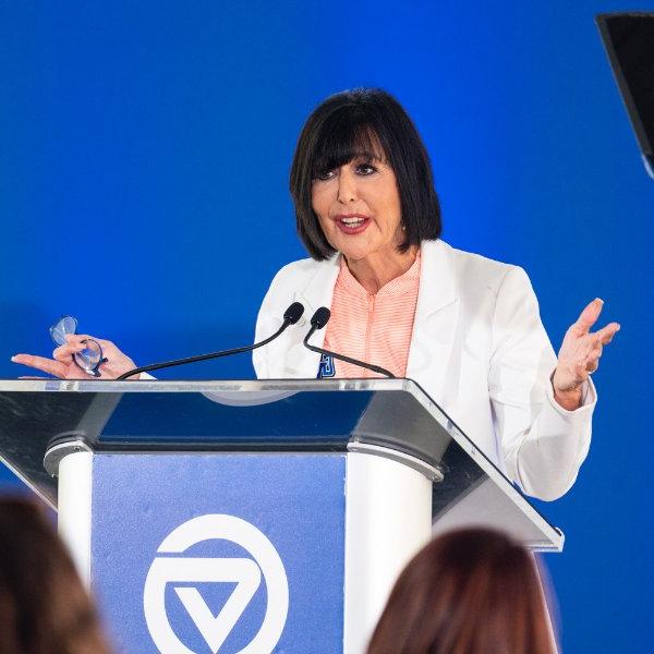 A person standing at a podium with the GVSU logo gestures with both hands while speaking. The person is holding eyeglasses in the left hand.