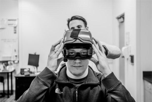 Justen Knape, a graduate student, dons an immersive headset in the Community Hearing Clinic, in Finkelstein Hall, with the help of faculty member Colton Clayton, who is standing behind him.